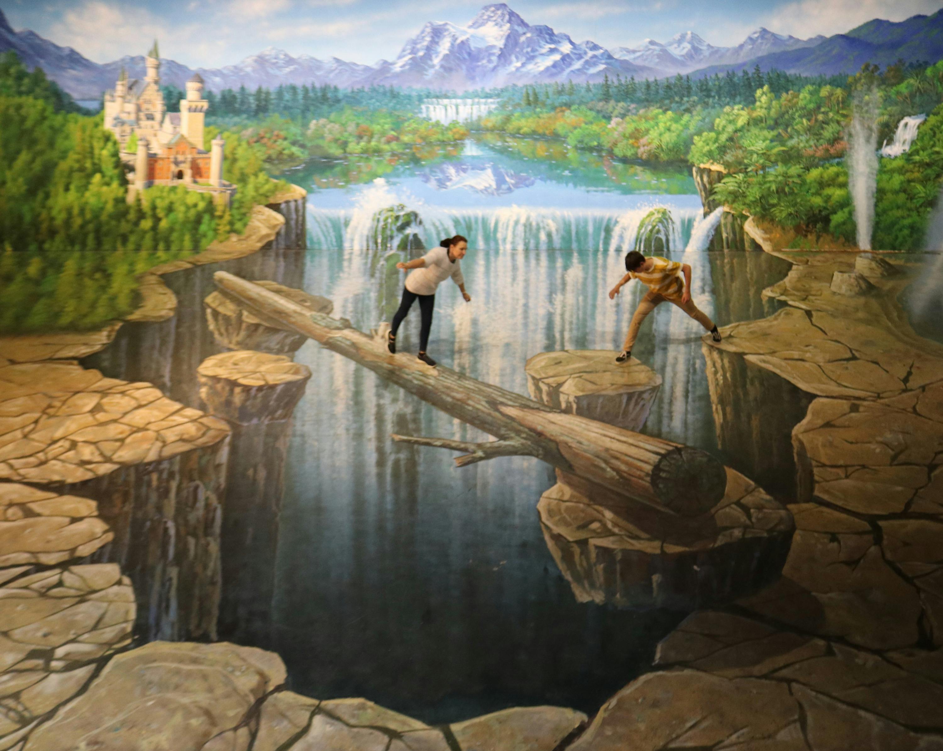 Two individuals interact with a painting that depicts a log bridge over a chasm, creating a playful optical illusion.