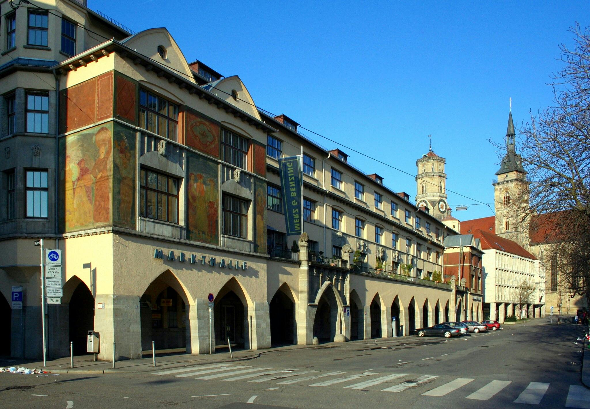 Street view of a multi-story market hall with arched windows, frescoes, and a "MARKTHALLE" sign, adjacent to historic buildings.