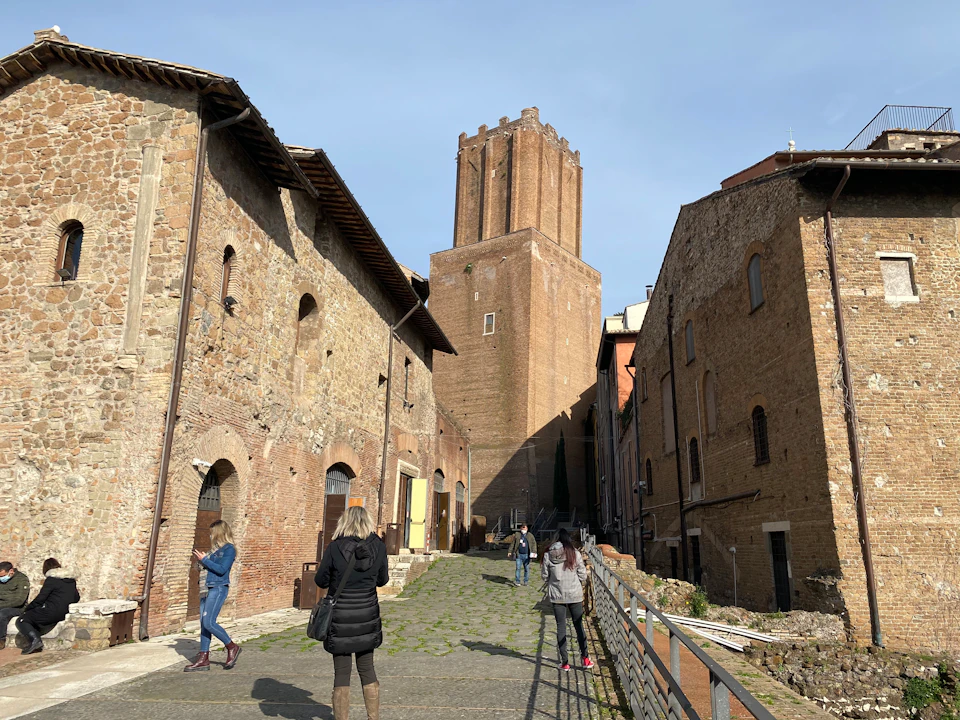 People walking along a cobblestone path flanked by historic brick buildings with a tall, square tower in the background.