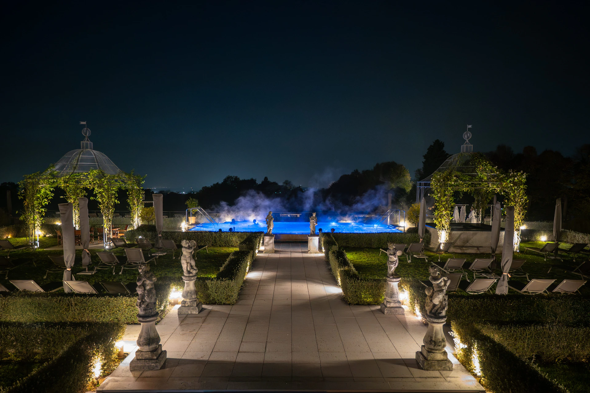 A luxurious outdoor pool area at night with illuminated statues, hedges, and a steaming blue-lit pool in the background.