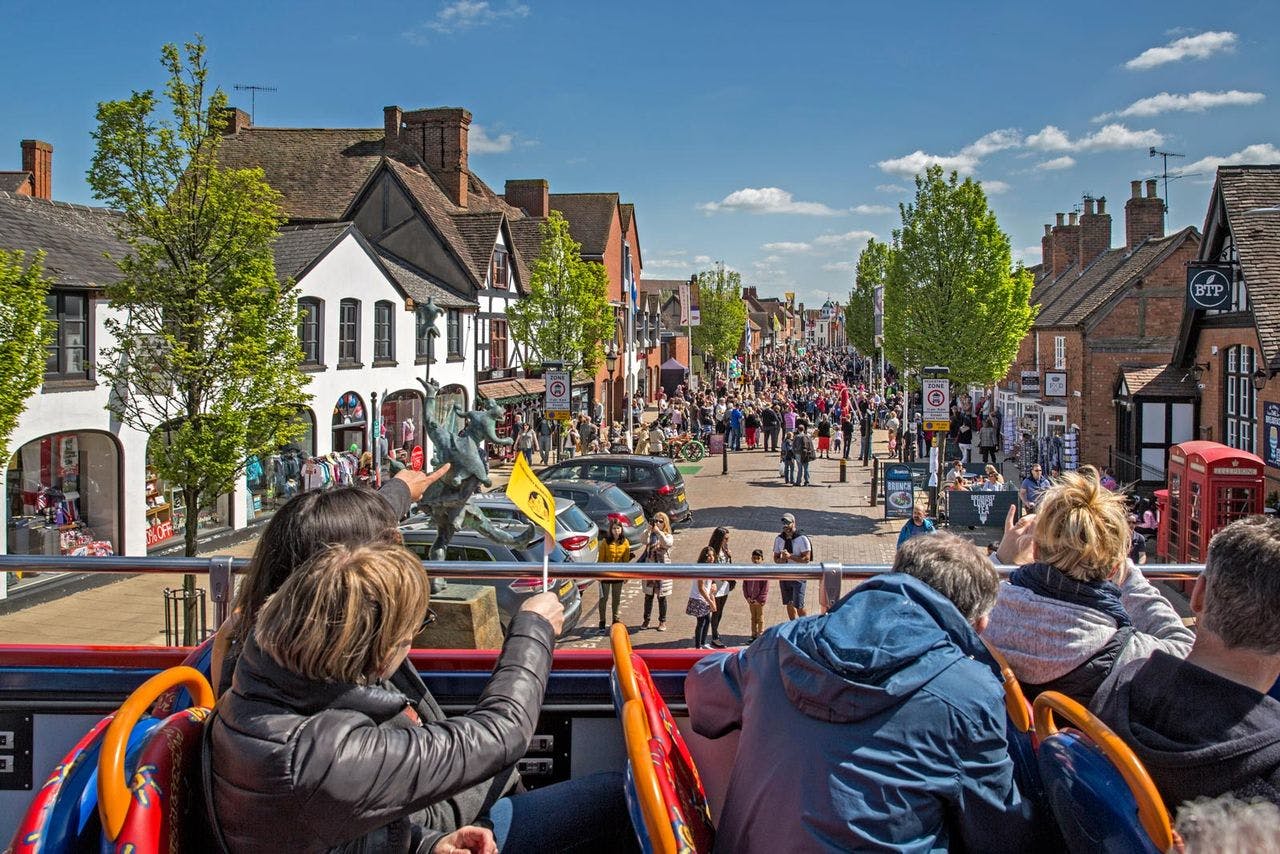 Rue bondée d'une ville animée, vue depuis un bus à toit ouvert. Les gens se promènent parmi les magasins et les arbres sous un ciel bleu lumineux.