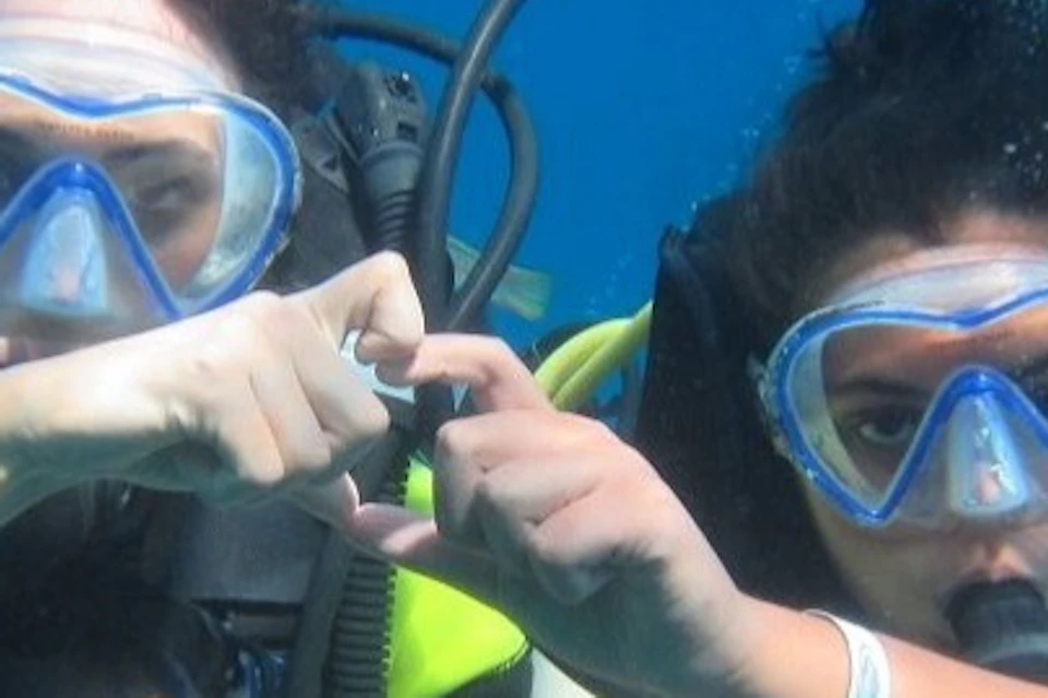 Two scuba divers underwater making hand signals, with a fish swimming nearby. Bubbles rise around them against the blue water.