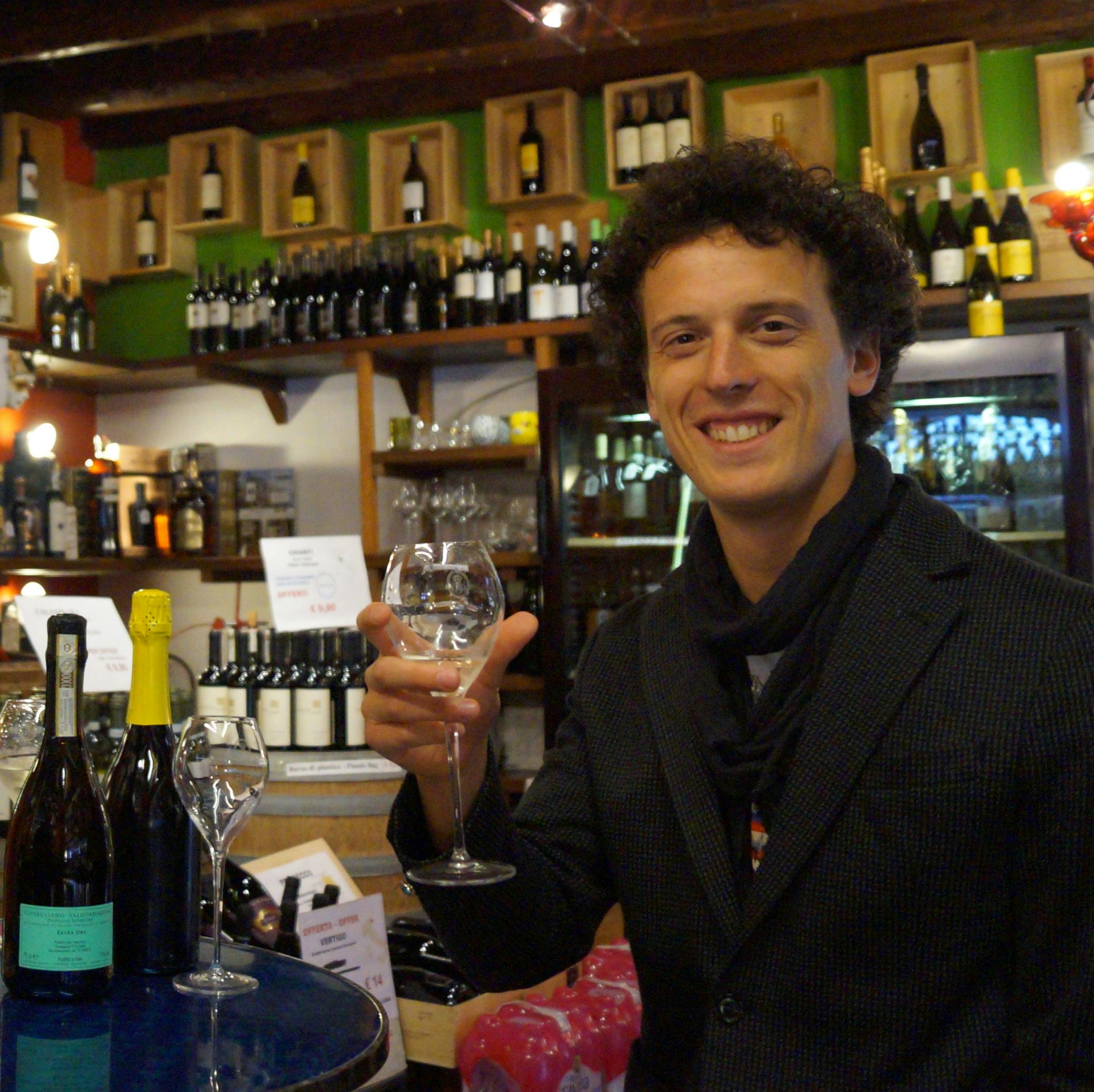 一名穿着西装的男子拿着葡萄酒杯，站在一家装满瓶子的葡萄酒商店的货架和柜台上。