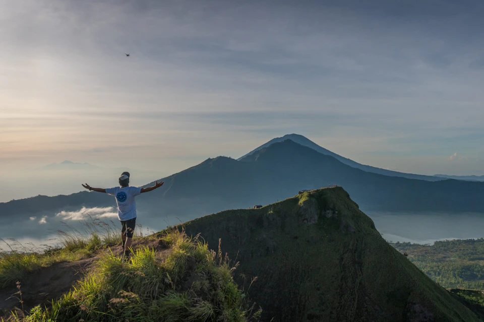 A person stands on a grassy mountain peak, arms outstretched, facing distant mountains under a clear sky.
