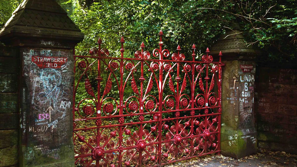 Ornate red wrought-iron gate with intricate patterns, set between stone pillars, surrounded by dense greenery.