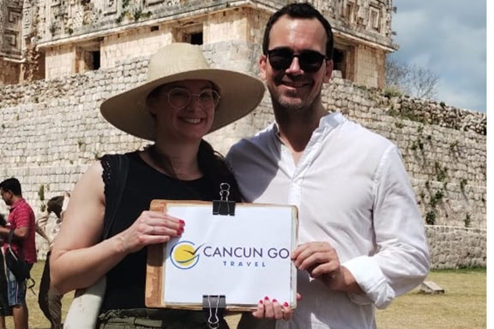 Deux personnes posant avec un panneau "Cancun Go Travel" devant un bâtiment historique en pierre. L'un porte un chapeau et des lunettes, l'autre des lunettes de soleil.