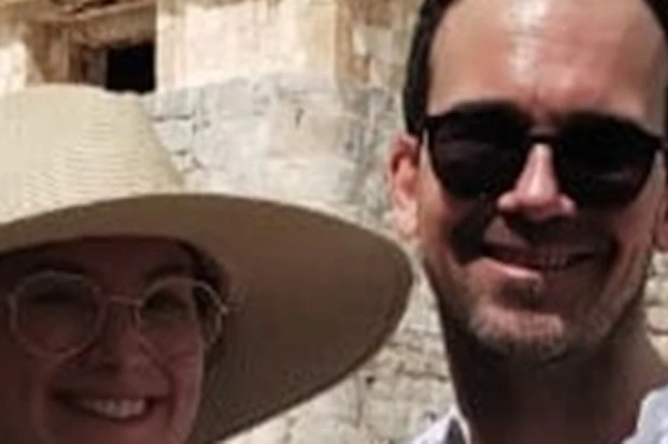 Two people posing with a "Cancun Go Travel" sign in front of a historic stone building. One wears a hat and glasses, the other sunglasses.