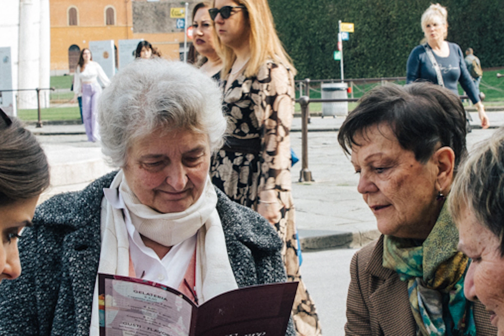 People sitting at an outdoor café table near the Leaning Tower of Pisa, with others walking and interacting in the background.