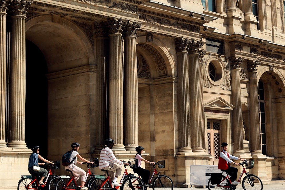 Four people ride red rental bicycles past an ornate building with large stone columns.