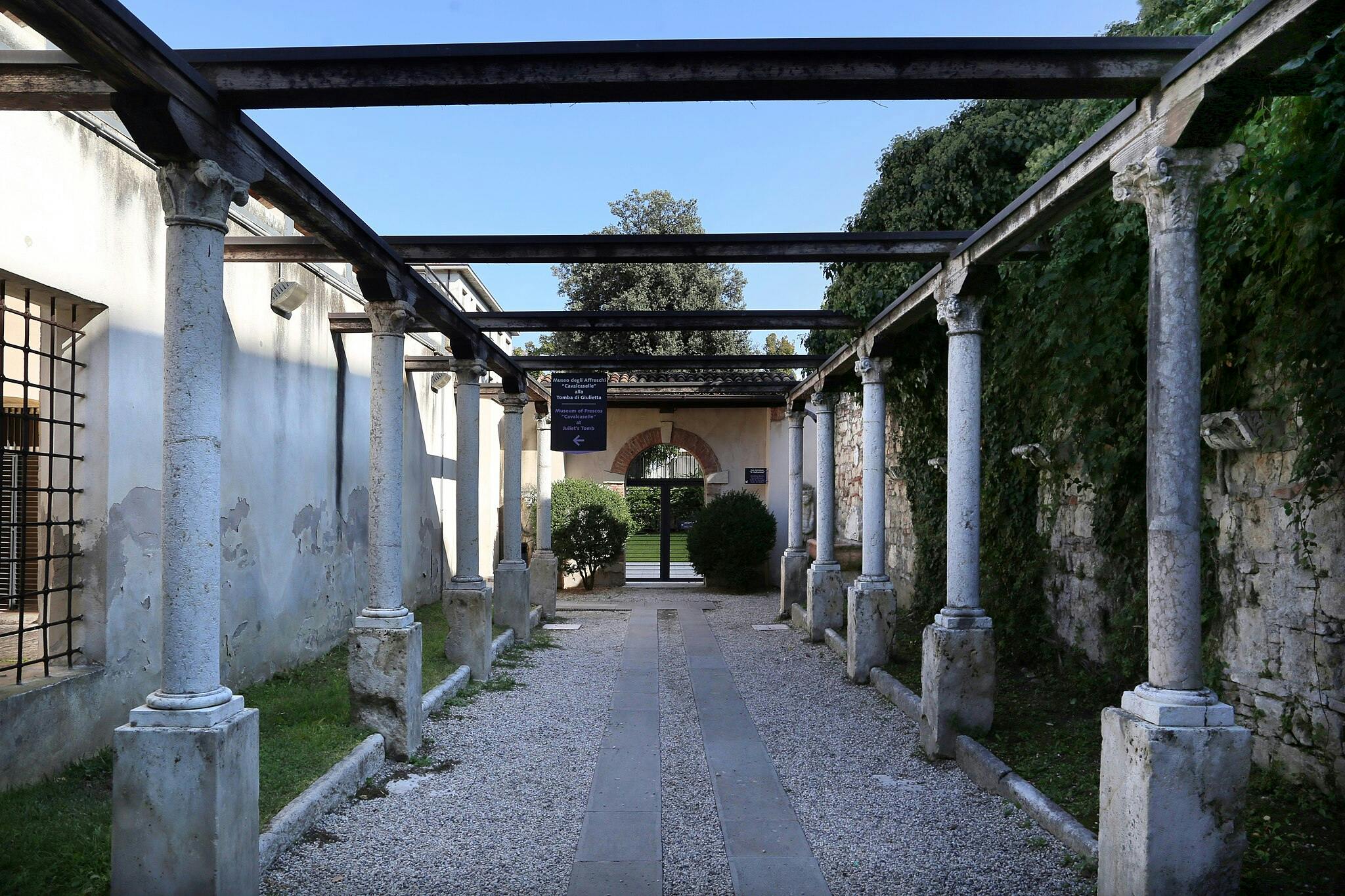 Le colonne di pietra sostengono le travi aeree lungo un sentiero di ghiaia, che conduce a un arco con il verde al di là.