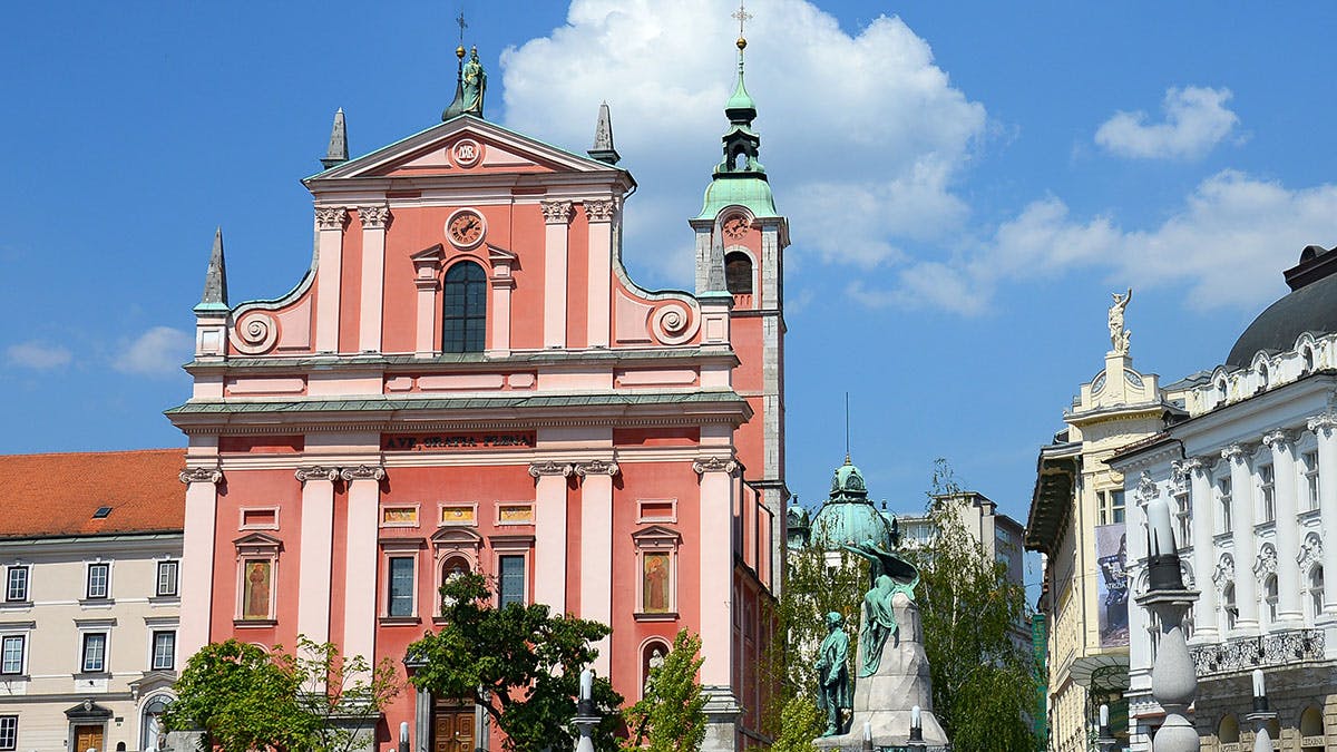 Růžová barokní fasáda kostela se zvonicí, sochou vpředu, stromy a modrou oblohou s mraky v pozadí.