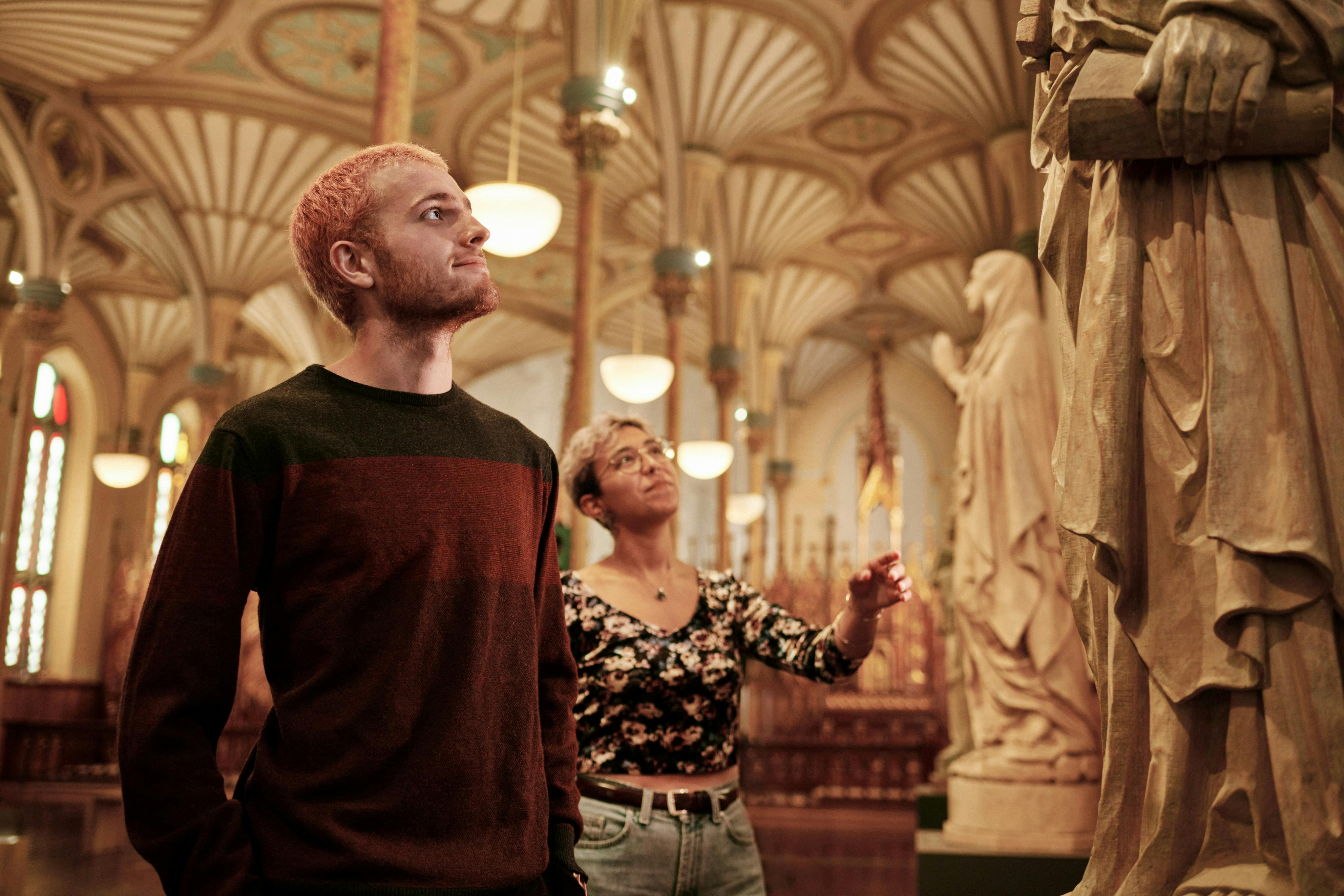 Two people with short hair admire the interior of a grand building with ornate columns and decorative ceiling patterns.