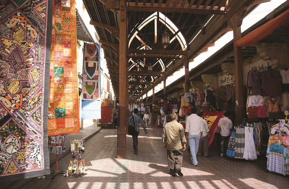 People walk through a busy, covered market lined with colorful textiles and various goods on display under wooden arches.