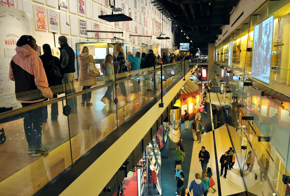 People walk along a two-story interior space with glass railings, several displays, and framed posters on the walls.