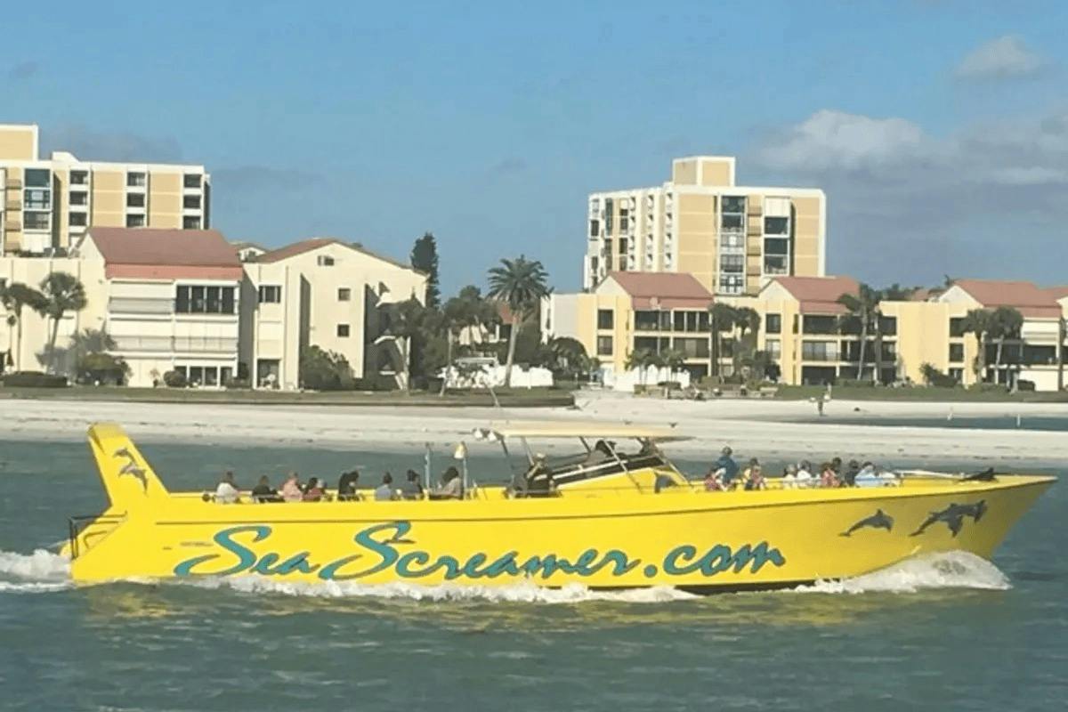 A bright yellow boat with "SeaScreamer.com" on its side is sailing in front of a beach with buildings in the background.