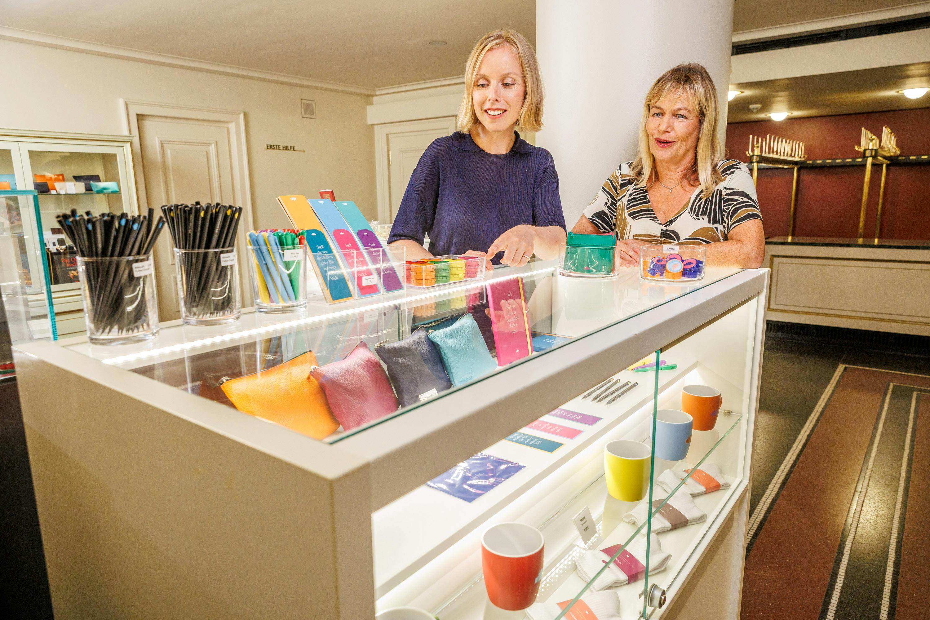 Two women examining colorful items displayed in a glass showcase at a well-lit retail setting.