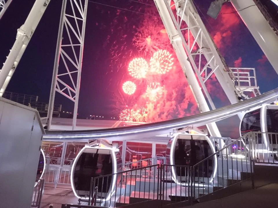 Fireworks explode in a night sky, viewed from within the structure of a large Ferris wheel, with gondolas in the foreground.