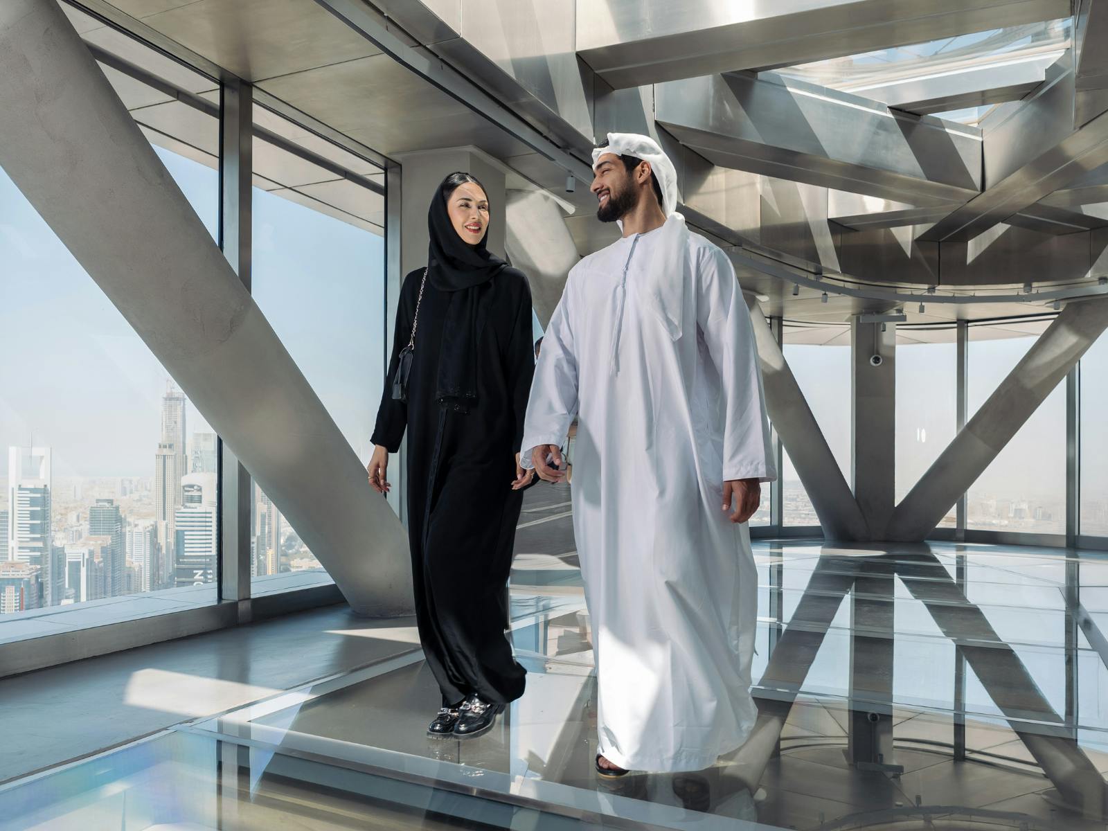 Muž a žena v tradičním oděvu vejdou ruku v ruce do moderní prosklené budovy s výhledem na město.