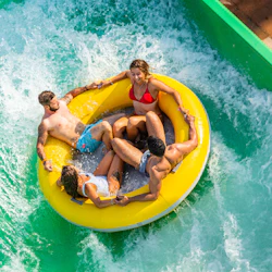 Four people riding an inflatable yellow raft down a water slide surrounded by green sides and splashing water.
