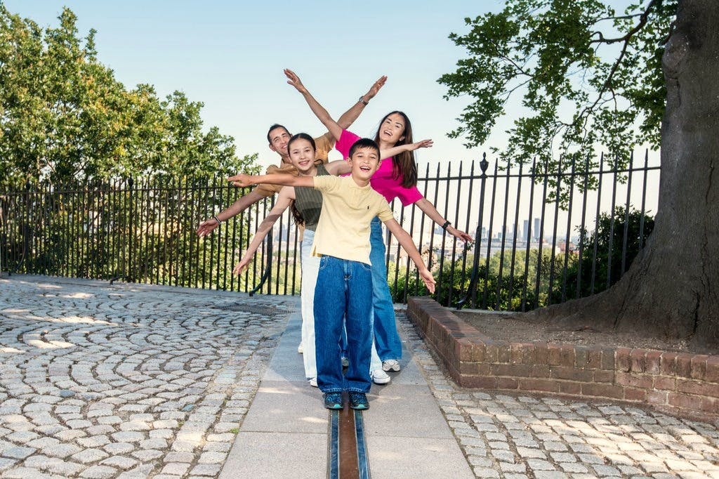 Cztery osoby stoją w kolejce z wyciągniętymi ramionami, uśmiechnięte i pozujące przy ogrodzonym terenie w parku z brukowanymi ścieżkami i drzewami.