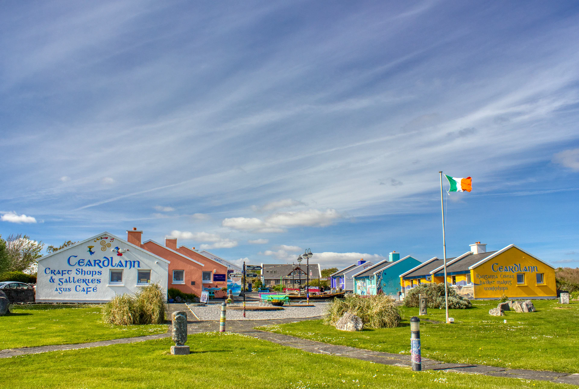 Colorful small buildings with a playground, Irish flag on a pole, and grassy park under a partly cloudy sky.