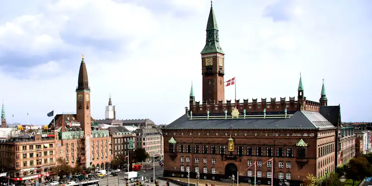 En stor, utsmyckad byggnad med torn och en klocka, danska flaggor, livlig gata med bussar och människor, mulen himmel.