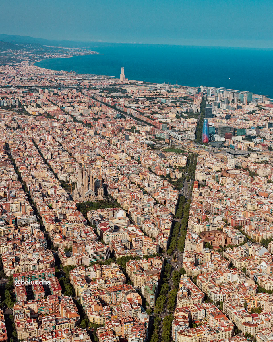 Aerial view of a densely packed city with a mix of old and modern buildings, a prominent cathedral, and the sea in the background.