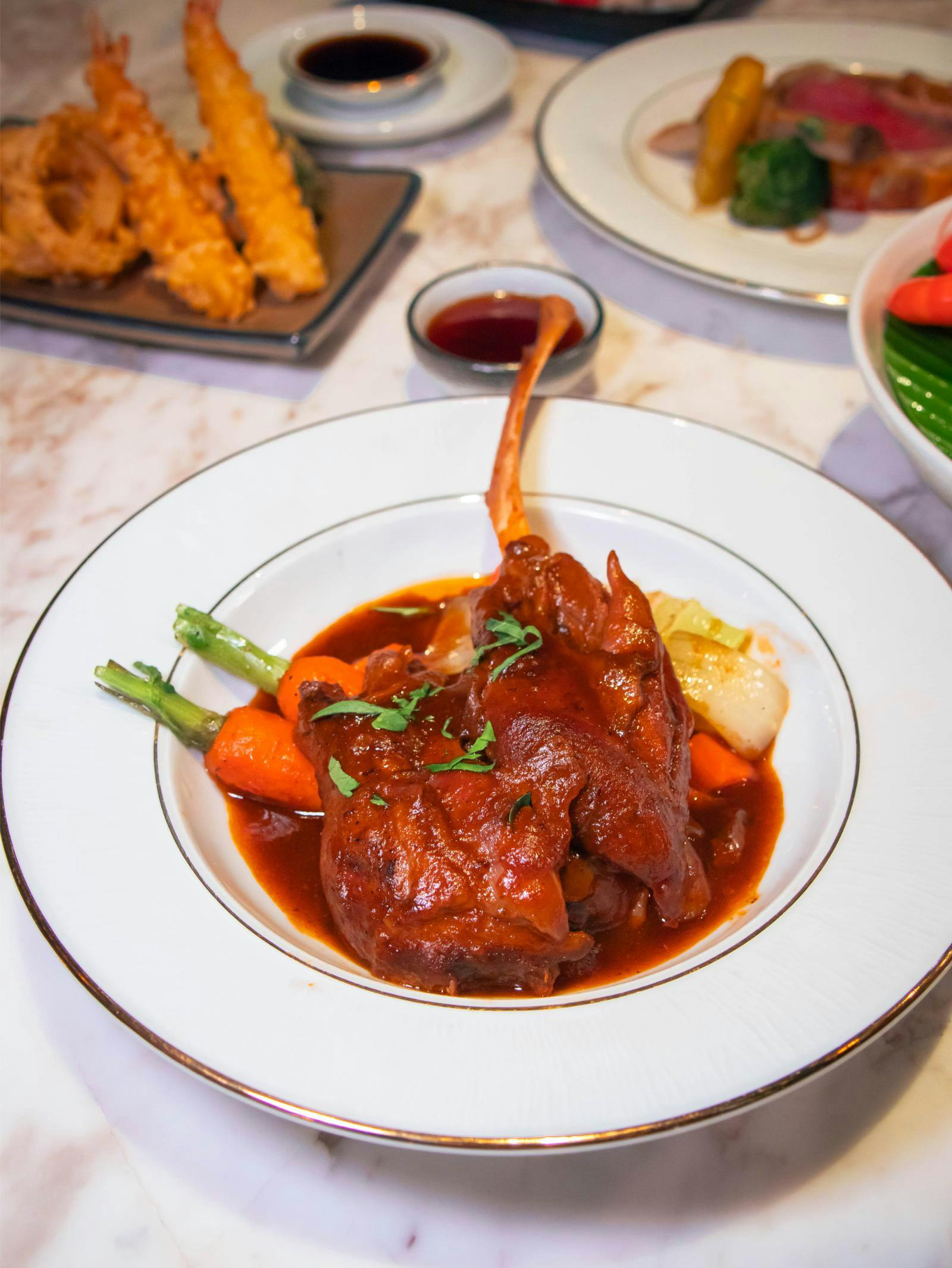 A plated dish featuring braised lamb shank with vegetables in a rich sauce, garnished with herbs.