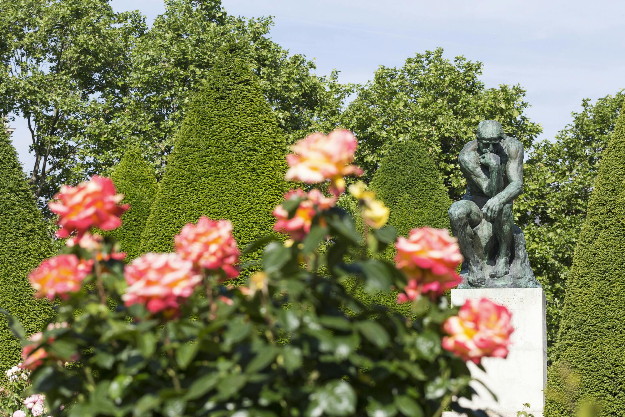 A garden with pink flowers in the foreground and a bronze sculpture of a thinking figure in front of neatly trimmed hedges.