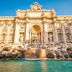 Utsmyckad fontän med statyer, kaskadvatten och en storslagen arkitektonisk bakgrund mot en klarblå himmel.