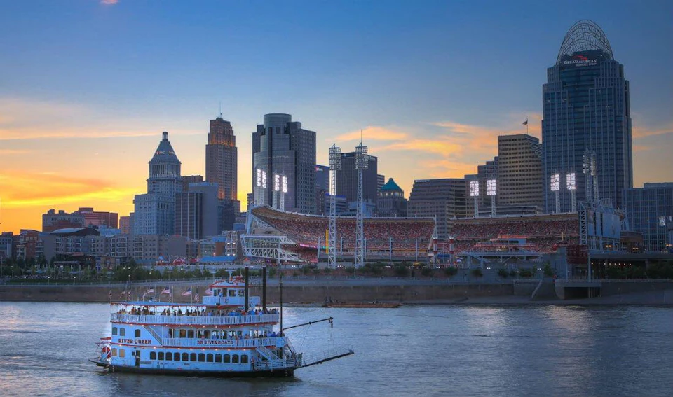 Skyline view of a city at sunset with a riverboat in the foreground and a sports stadium illuminated in the background.