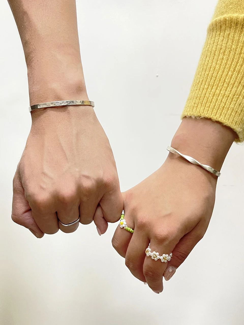 Two hands with bracelets and rings, one wearing a yellow sleeve and the other bare, touching lightly against a plain background.