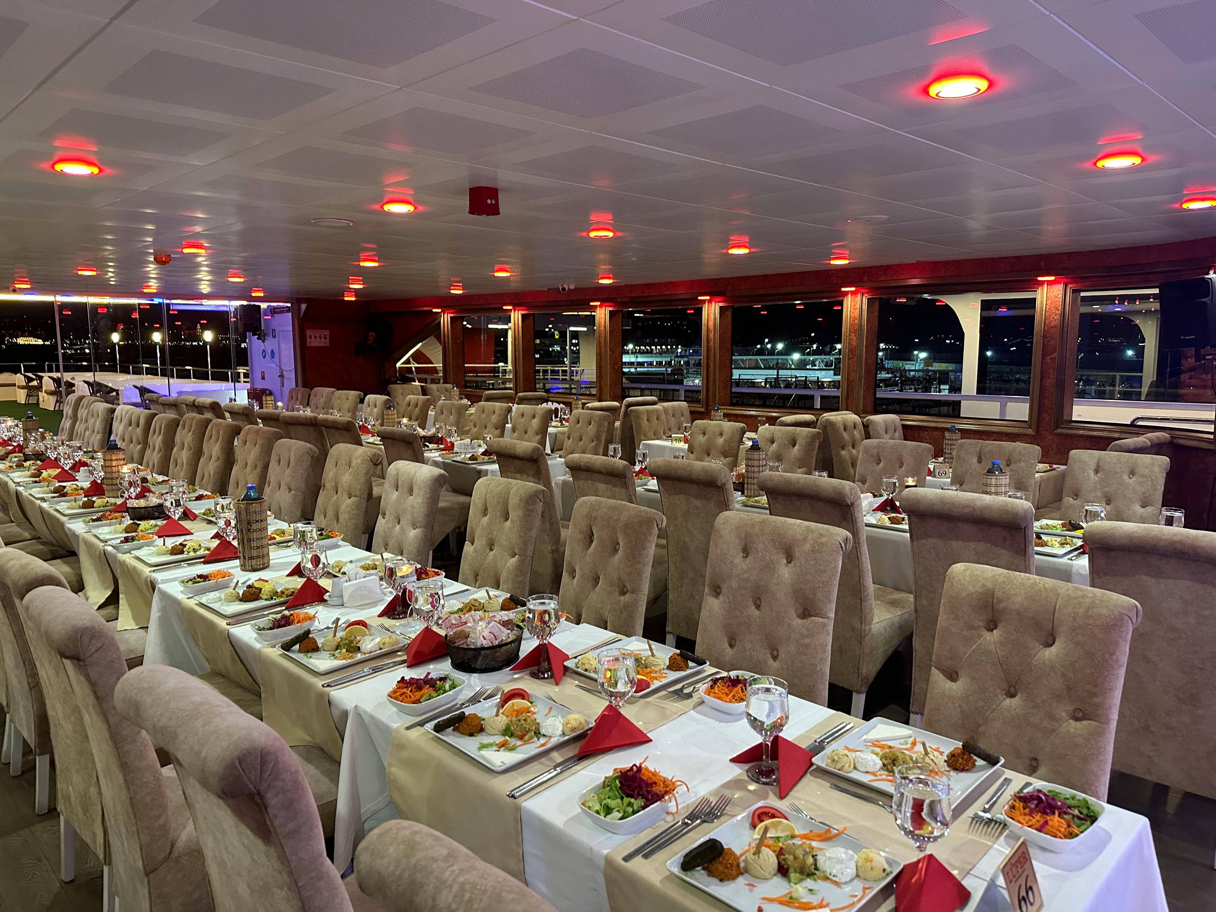 An elegant dining area on a ship with multiple tables set with plates, cutlery, and red napkins, overlooking city lights.