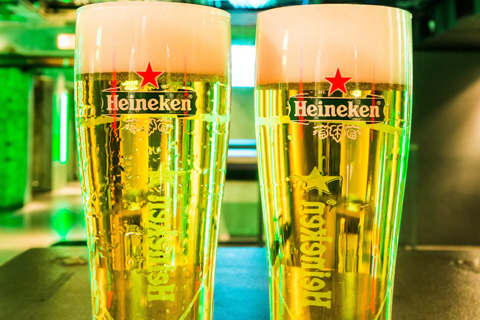 Two glasses of beer with Heineken branding, filled with golden liquid and foam on top, placed on a bar surface.