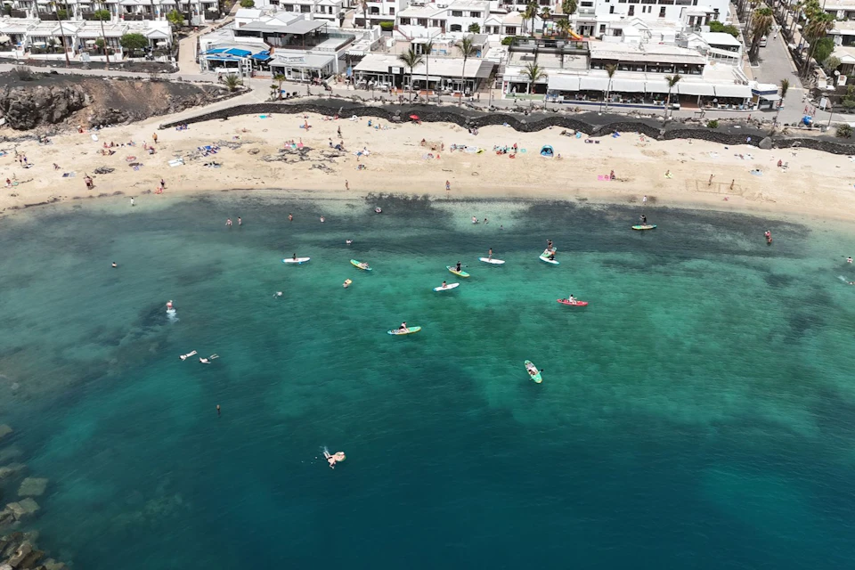 People paddleboarding and swimming in clear turquoise water near a sandy beach, with white buildings in the background.