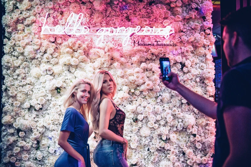 Two people posing in front of a floral wall with a neon sign reading "hello gorgeous" and "Madame Tussauds."