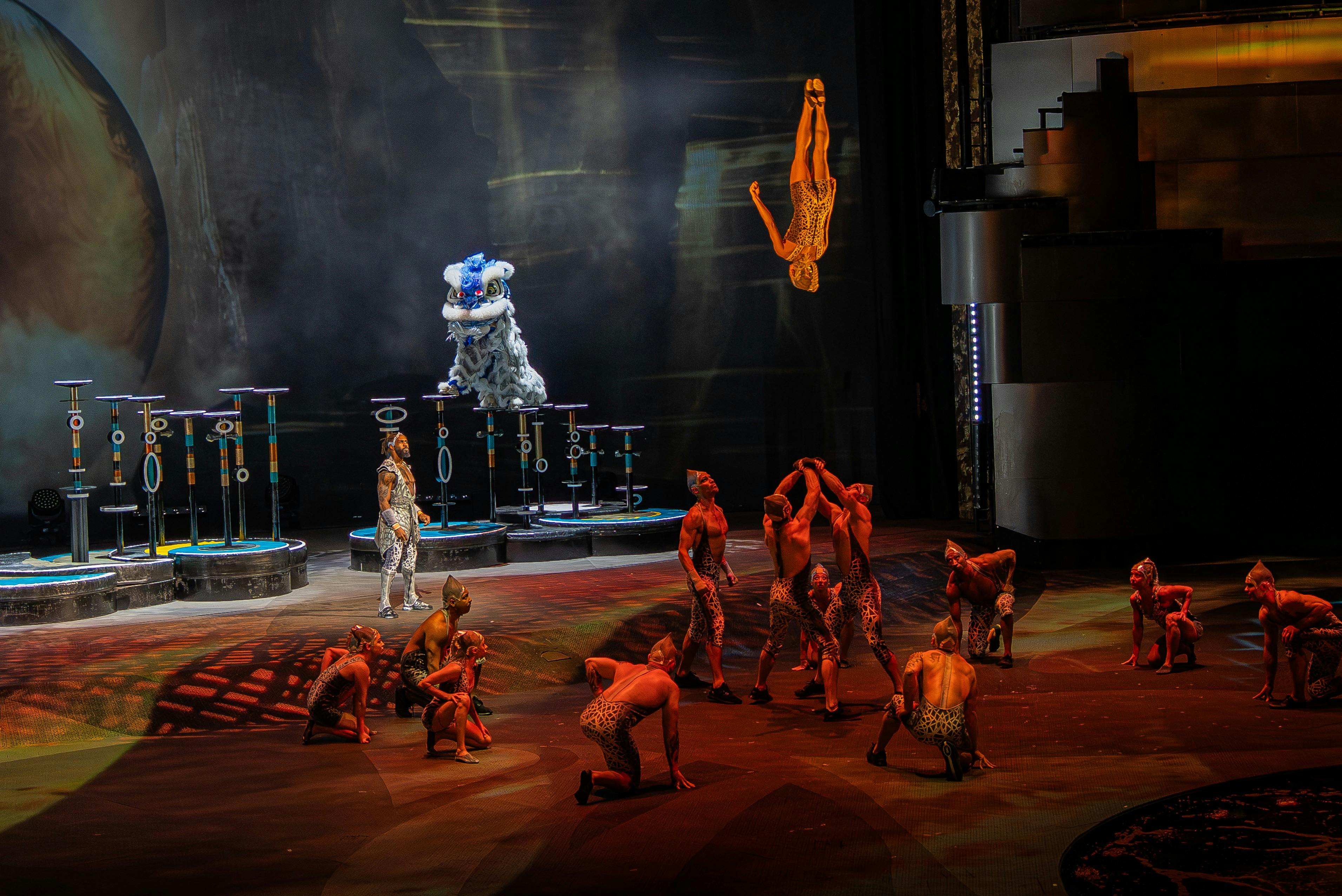 Uitvoerenden op het podium in een acrobatische act, met één persoon in de lucht en anderen balancerend of poserend onder de podiumlichten.