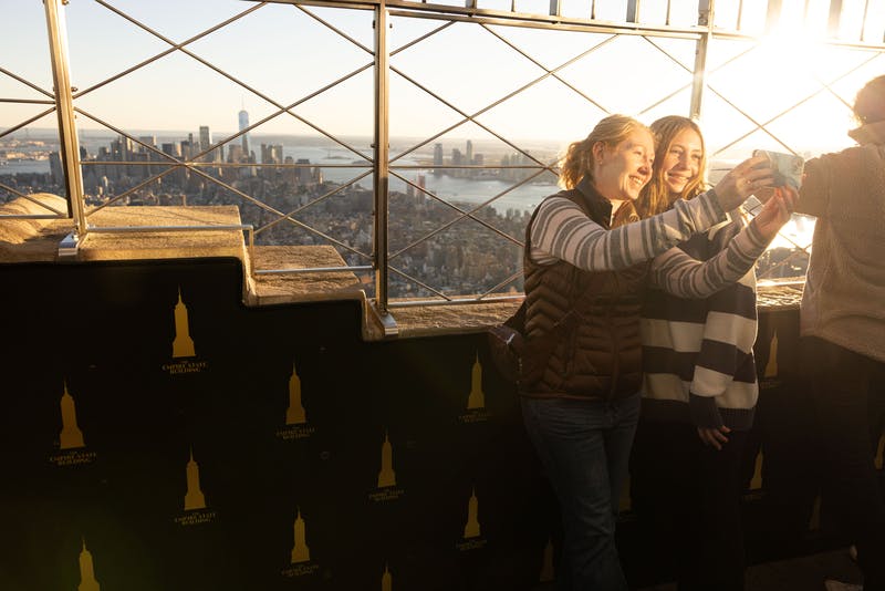 Δύο άνθρωποι χαμογελούν και βγάζουν μια selfie σε ένα ψηλό κατάστρωμα παρατήρησης, με φόντο τον ορίζοντα της πόλης και ένα ποτάμι.