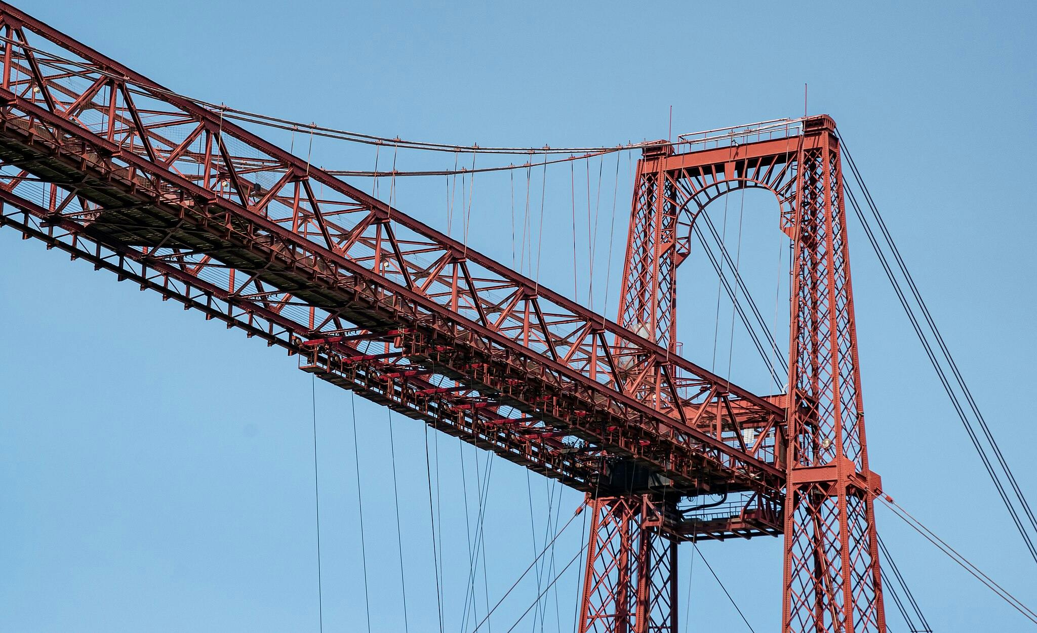 Close-up of a red steel suspension bridge's tower and cables against a clear blue sky.
