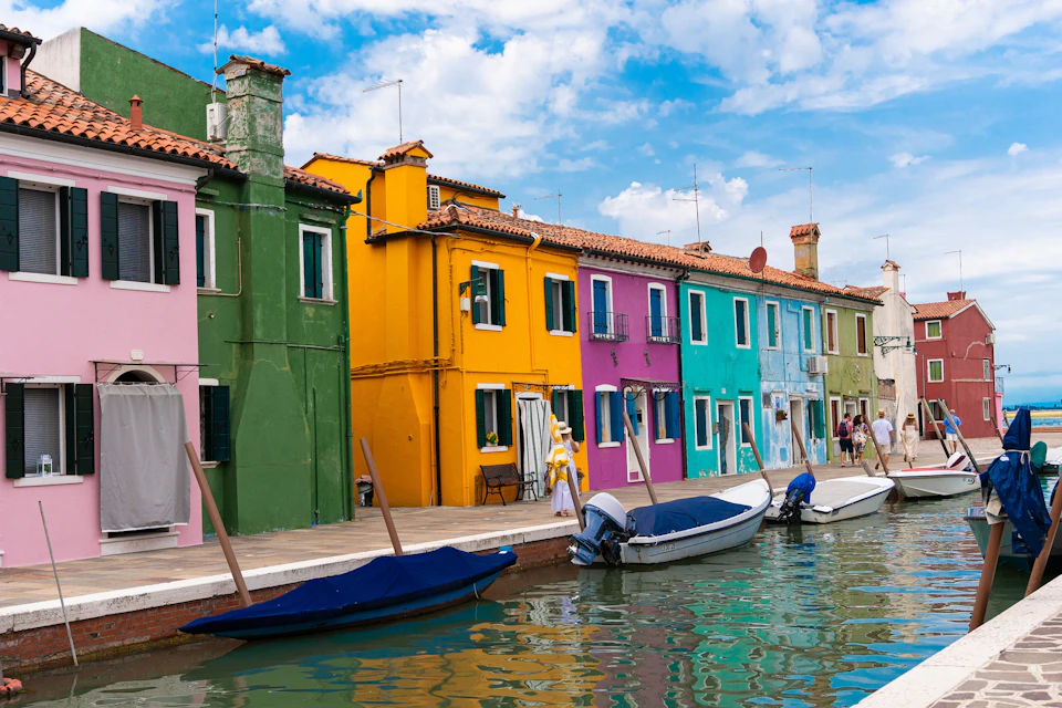 Brightly colored houses line a canal with moored boats. People walk along the waterfront under a partly cloudy sky.