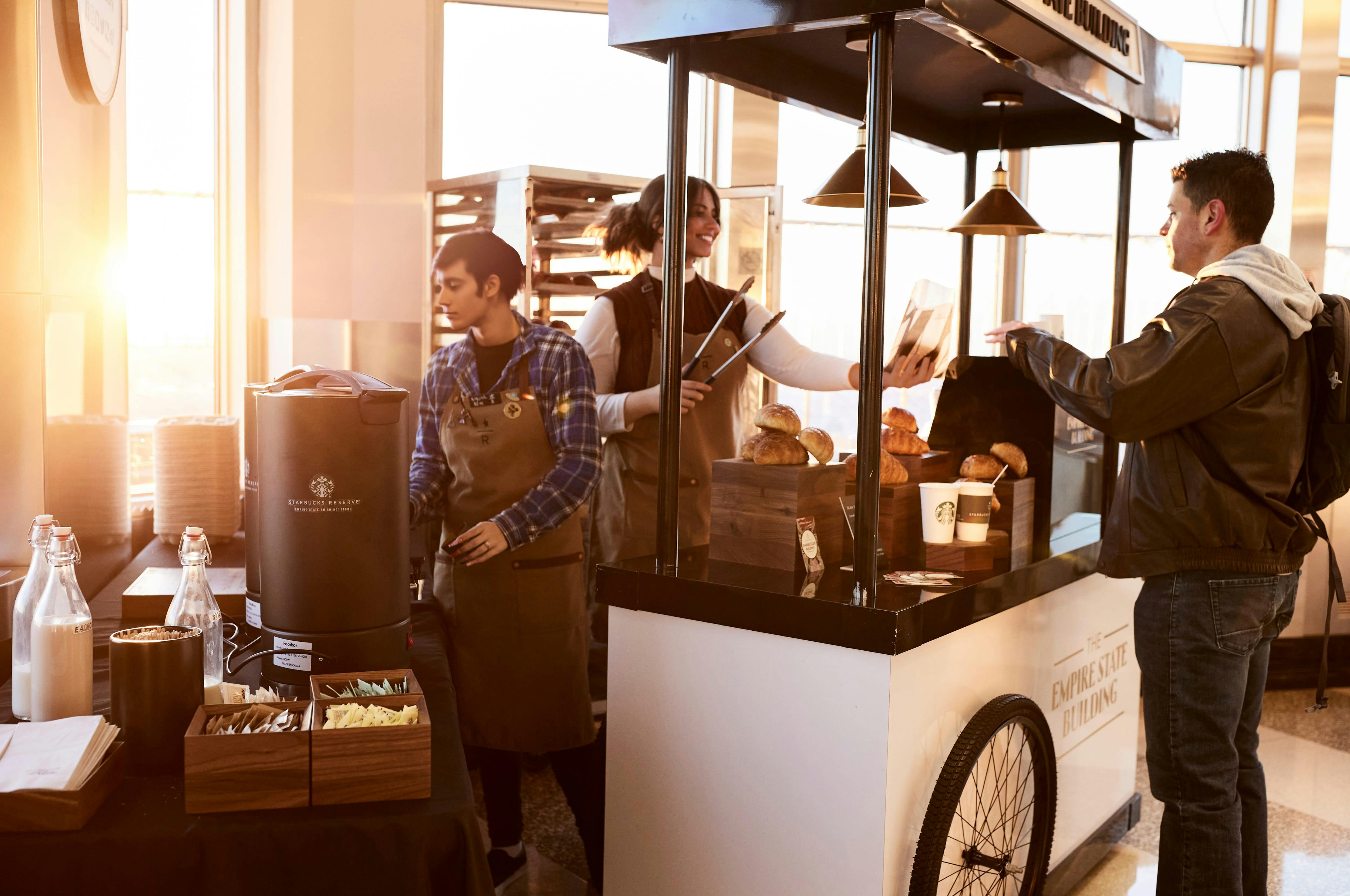 Two bakery workers in aprons prepare items on a counter with baked goods and drinks, illuminated by warm sunlight.