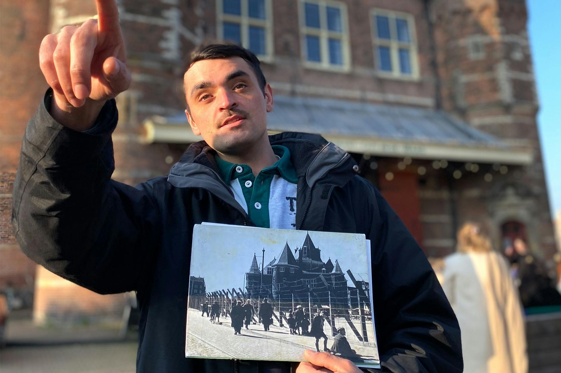 어두운 재킷을 입은 한 남성이 창문이 있는 벽돌 건물 앞에 서서 가리키면서 역사적인 사진을 들고 있습니다.