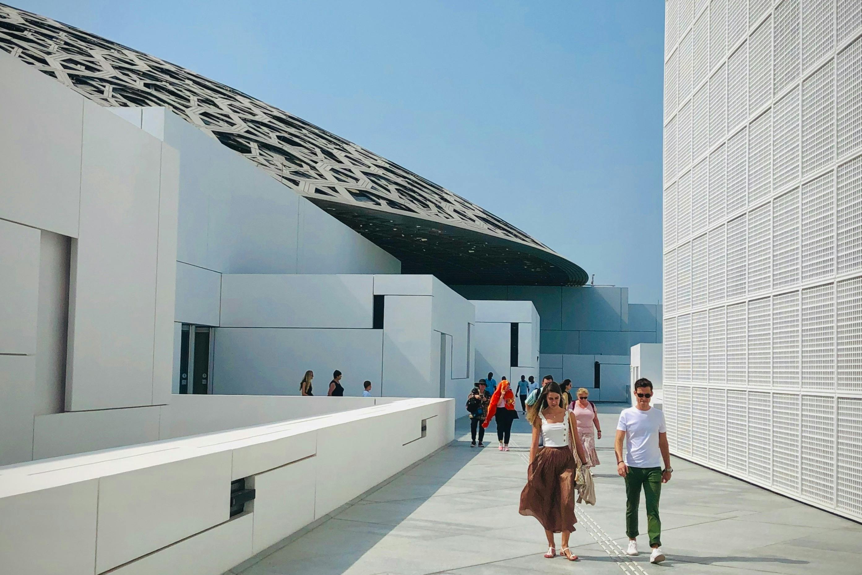 People walking on a modern architectural pathway with white walls and a patterned dome structure visible in the background.