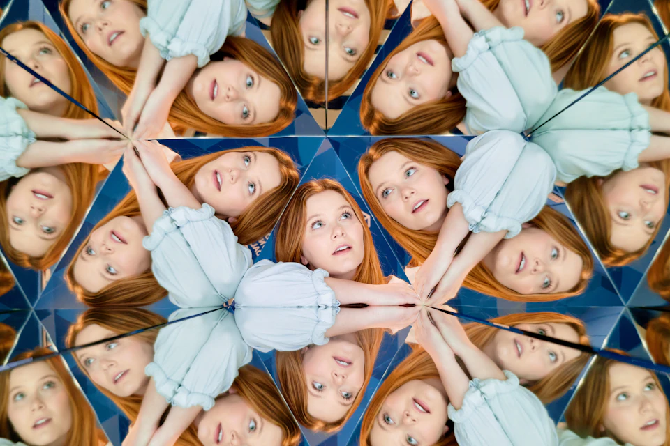 A woman with red hair and a light blue blouse is reflected multiple times in a kaleidoscope-like mirror pattern.