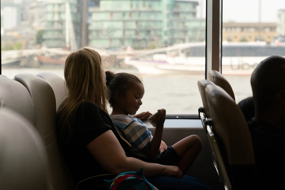 A woman and a child sit together on a boat with a view of a river and buildings through the window.