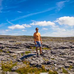 En person i solglasögon och lediga kläder står på en klippa under en klarblå himmel med spridda moln.