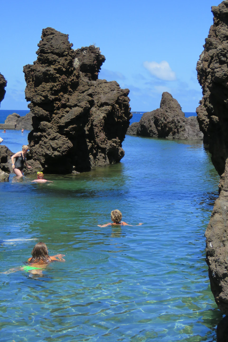 People swimming in a clear, rocky tide pool surrounded by large rock formations under a blue sky.