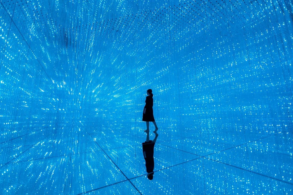 A person stands in a mirrored room filled with blue lights, creating the illusion of endless light rays.