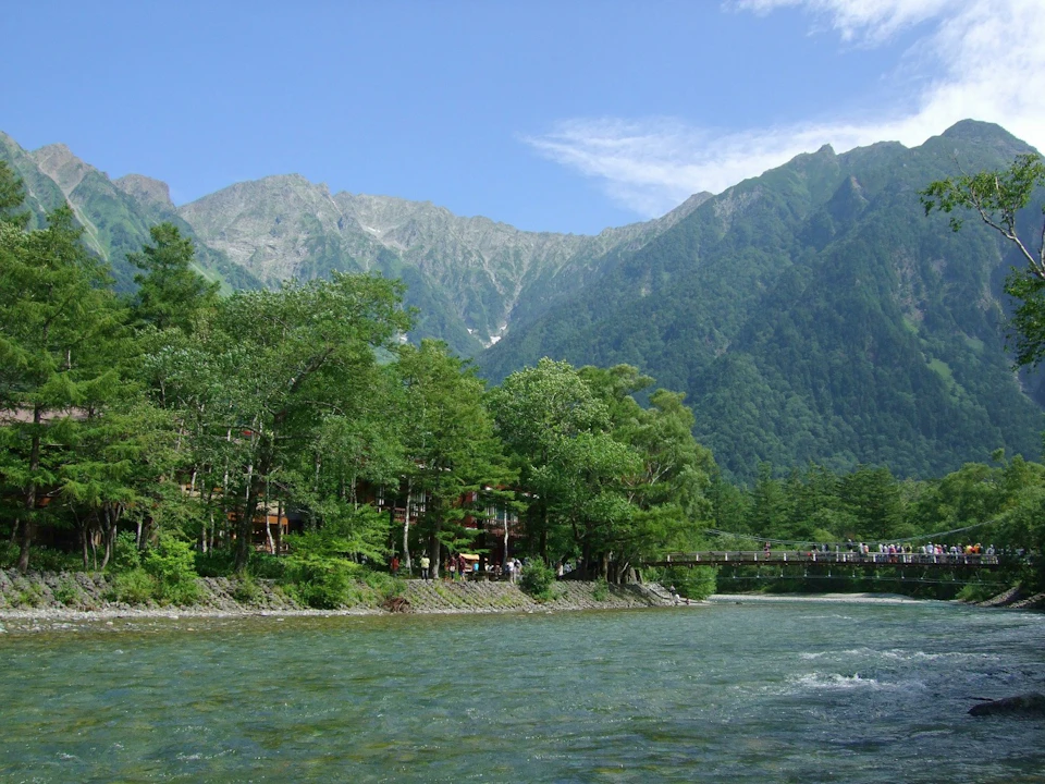 A scenic river flows with a forested area and a bridge in the background, set against a mountainous backdrop under a blue sky.
