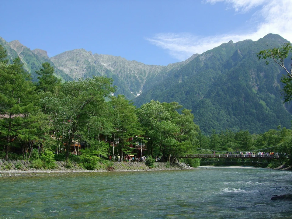 A scenic view of a river with a tree-lined shore, a bridge, and mountains in the background under a blue sky.